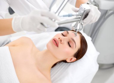 Modern güzellik kliniğindeki kadın müşterinin yüzüne lazer teçhizatı kullanan profesyonel bir kozmetikçi.