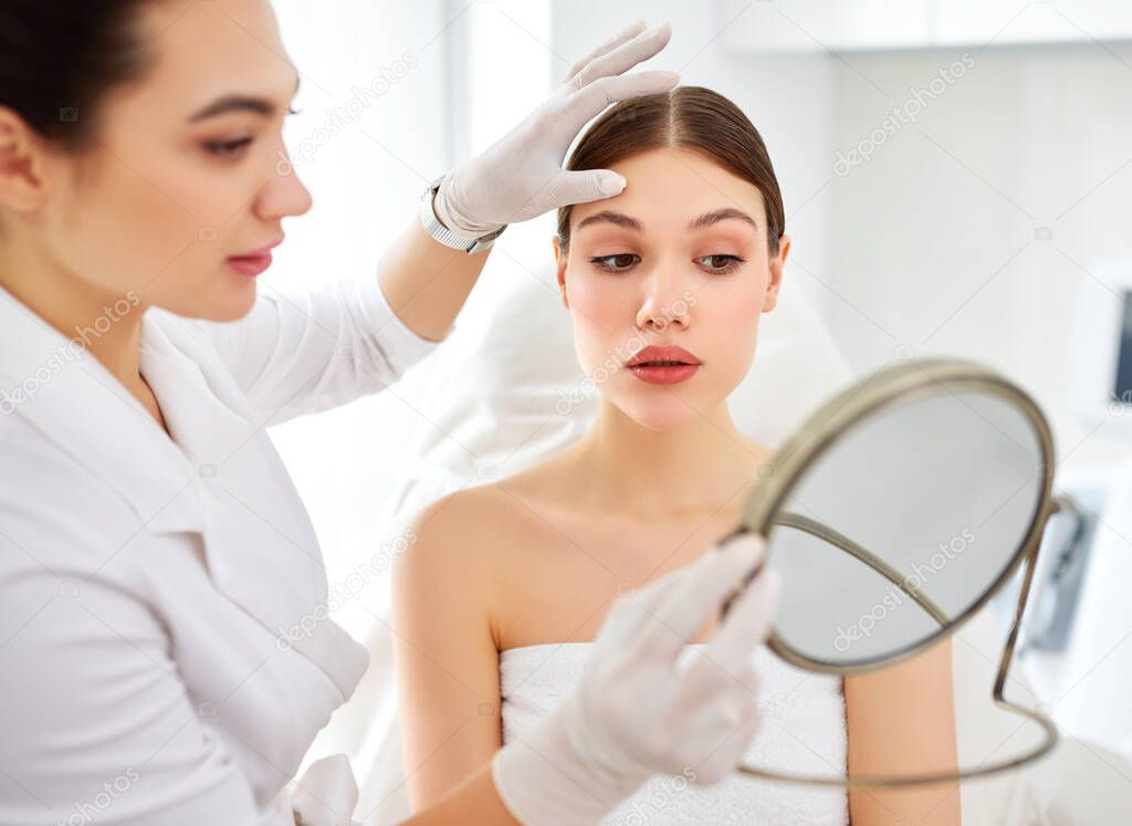 Satisfied young woman looking at mirror and touching face after professional cosmetology treatment with beautician in modern beauty clinic