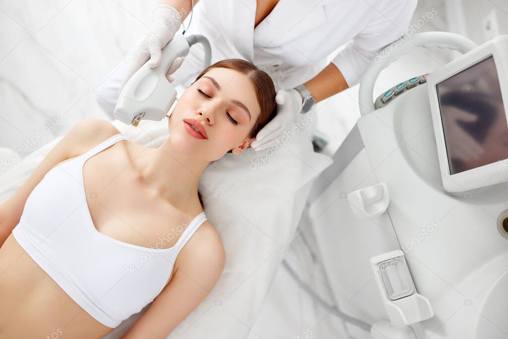From above of young female client receiving laser hair removal procedure our receiving ultrasound face lifting during rejuvenation procedure in beauty clinic on face during beauty treatment in modern salon