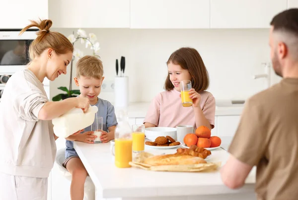 年轻而积极的妈妈在和两个可爱的孩子 女儿和儿子 一起吃着健康的早餐的同时 从玻璃瓶里倒入新鲜牛奶 一家四口快乐地在厨房里吃着早餐 — 图库照片