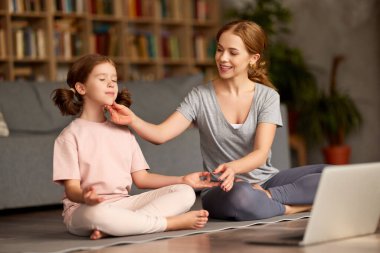 Genç pozitif anne, Lotus 'ta birlikte oturma odasında poz verirken ve dizüstü bilgisayardan yoga dersleri izlerken tatlı küçük kızına nasıl meditasyon yapacağını gösteriyor.