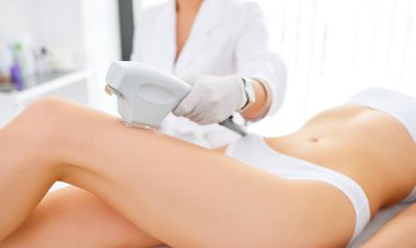 Tanımlanamayan profesyonel kozmetik uzmanı lazer aletli kadın hastanın bacaklarındaki kılları modern güzellik merkezinde lazer cilt bakımı sırasında alıyor.