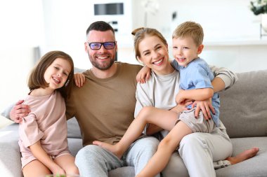Dört çocuklu mutlu bir ailenin portresi, baba, anne ve iki sevimli çocuk kanepede oturup kucaklaşırken kameraya gülümsüyorlar, çocuklu ebeveynler hafta sonlarını evde birlikte geçiriyorlar.