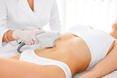 Modern salonda güzellik lazeri cilt bakımı sırasında bayan müşterinin bikini bölgesinde lazer tüy temizleme aparatı kullanan eldivenli isimsiz bir güzellik uzmanı.