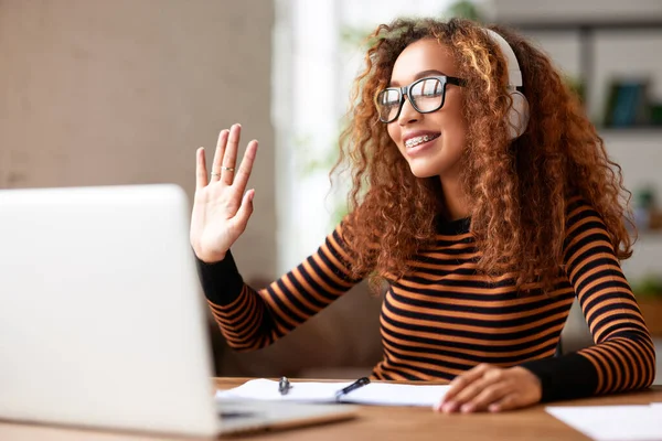 Cheerful young african american woman in headphones greeting colleague via webcam on laptop while working remotely from home, happy female student having video call with teacher while studying online