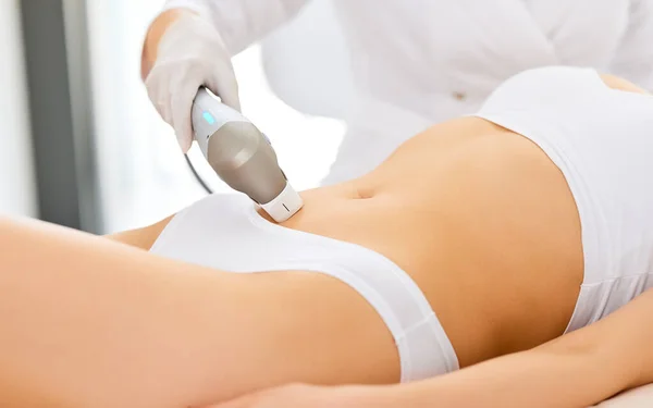 Crop anonymous beautician in gloves using laser hair removal apparatus on bikini zone of female client during beauty laser skin care procedure on stomach  in modern salon