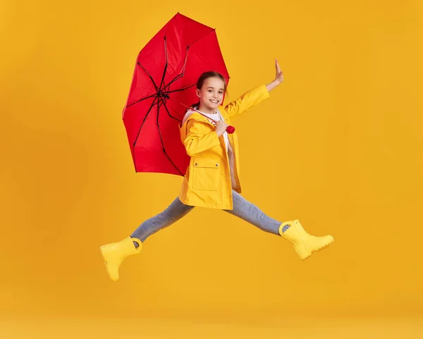 身穿黄色雨衣和红色雨伞靴子的兴奋小女孩的全身上下尽收眼底 在黄色背景下从地面上跳下 — 图库照片