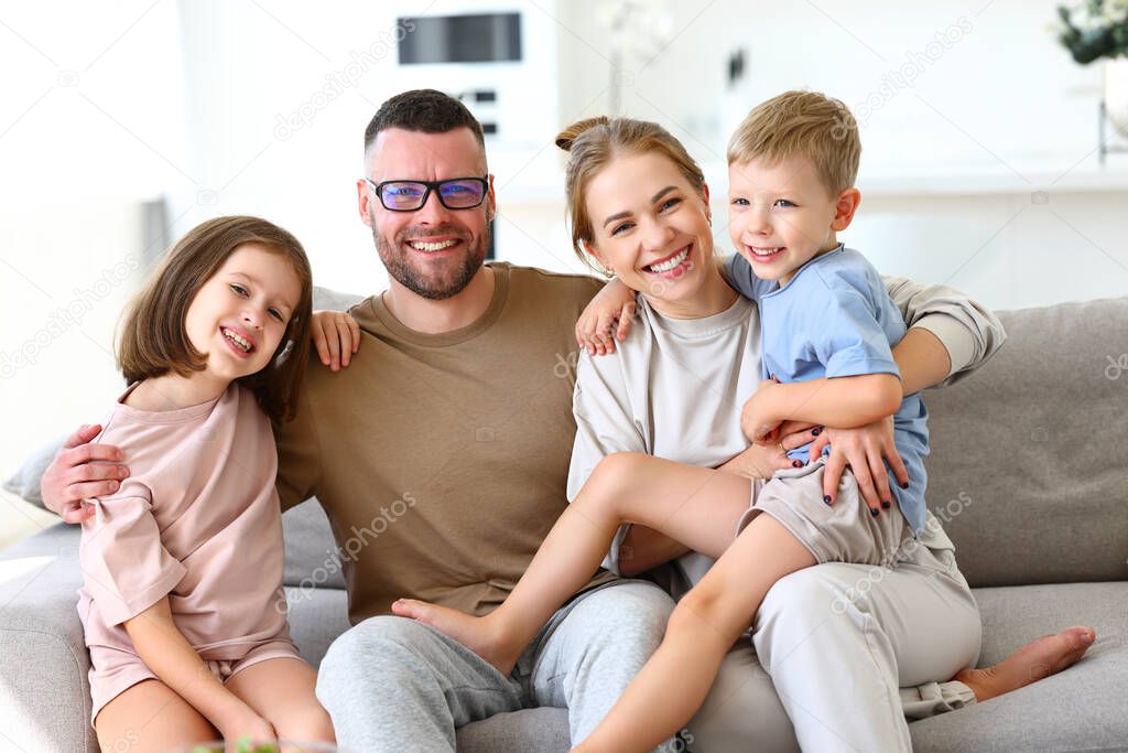 Portrait of beautiful positive happy family of four, father, mother and two cute children smiling at camera while sitting on sofa and embracing, parents with kids enjoying weekend together at home