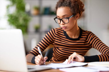 Odaklanmış genç Afro-Amerikan kadın bazı notlar alıyor ve iş yerinde oturup uzaktan çalışırken laptop ekranına bakıyor. Yoğunlaştırılmış melez kız öğrenci evde online çalışıyor.