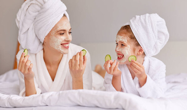 Young woman and girl with cucumbers smiling and looking at each other while relaxing on bed during spa procedure at home