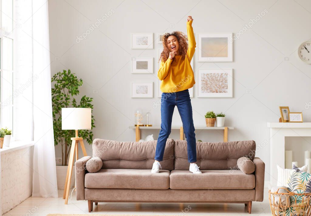 Pretending to be famous singer. Overjoyed afro american teenage girl in casual wear singing and dancing on sofa in modern living room at home. Young happy female enjoying carefree and leisure weekend
