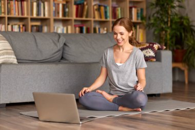 Spor elbiseli genç bir kadın dizüstü bilgisayarın önünde poz veriyor. Evde yoga yapan bir kadın. İnternetten ekrana bakıyor ve gülümsüyor.