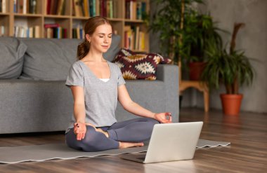 Spor giyim sektöründe genç, huzurlu bir bayan, gözleri kapalı meditasyon yapıyor, evde dizüstü bilgisayarında yoga yaparken nefes egzersizleri yapıyor, tam boy. Karantina sırasında eğlenen dikkatli kadın