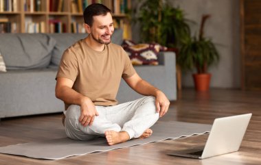 Gündelik giyinen genç adam yoga dersini internetten alıyor, dizüstü bilgisayara bakıyor ve Lotus 'ta poz verirken gülümsüyor. Covid19 karantinası sırasında spor