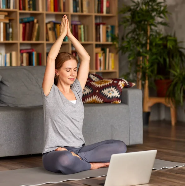 年轻和平的女性穿着运动服 闭着眼睛沉思 做呼吸练习 同时在家里的笔记本电脑上开设瑜伽课 全身而退 在检疫期间享受活动的妇女 — 图库照片