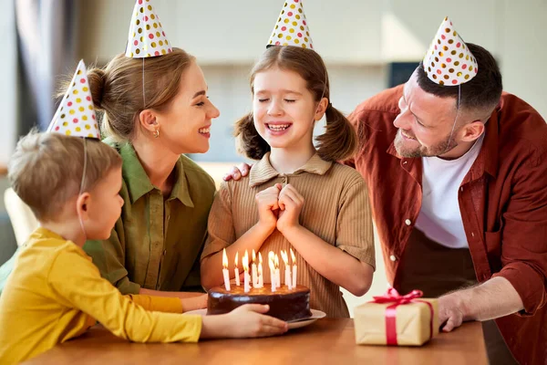 かわいい女の子はパーティー帽子をかぶって手で目を覆い 自宅で家族と誕生日を祝いながら願いを作り チョコレートケーキにキャンドルを吹き 両親は娘にギフトボックスを与えます — ストック写真