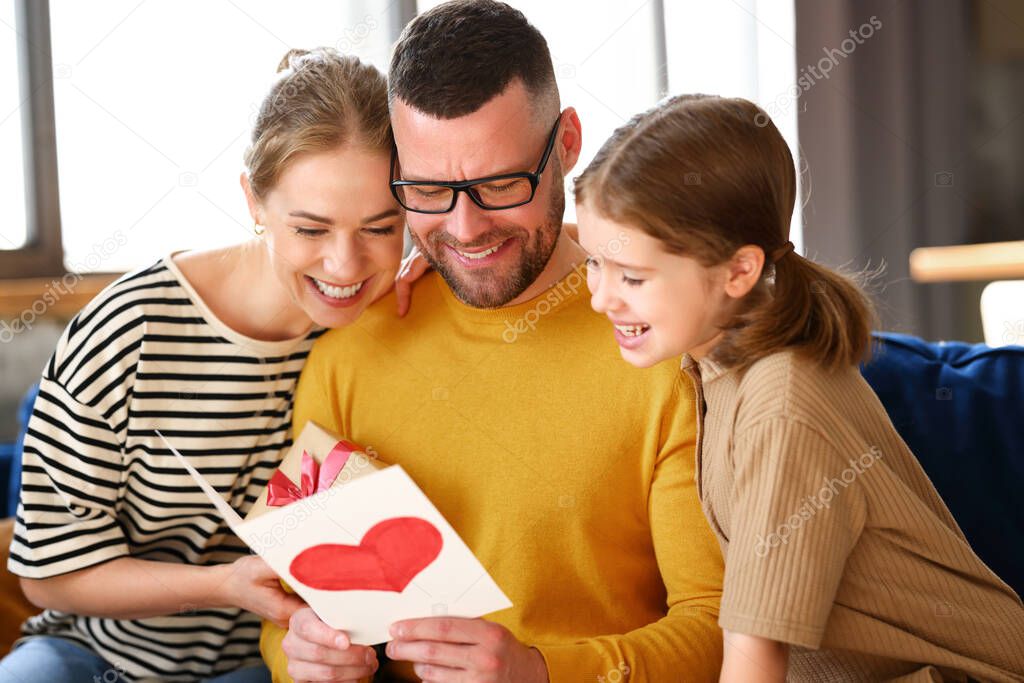 For best husband and dad. Happy family mother and little daughter congratulating father with holiday, giving him presents handmade postcard and wrapped gift box while sitting together on sofa at home