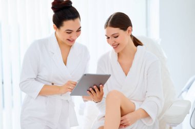 Modern güzellik kliniğindeki randevu sırasında cilt bakımı prosedürünü açıklarken gülümseyen kadın hastaya tablet gösteren pozitif profesyonel kozmetik uzmanı