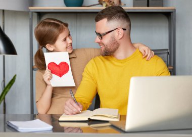 Küçük bir kız çocuğu, dizüstü bilgisayarda çalışırken babaya el yapımı kartpostal atıyor. Genç ve mutlu baba, evdeki çocuktan tebrik alıyor. Aile tatili konsepti