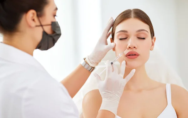 Professional beautician in protective mask and gloves applying injection on face of young female client during cosmetology treatment in beauty clinic