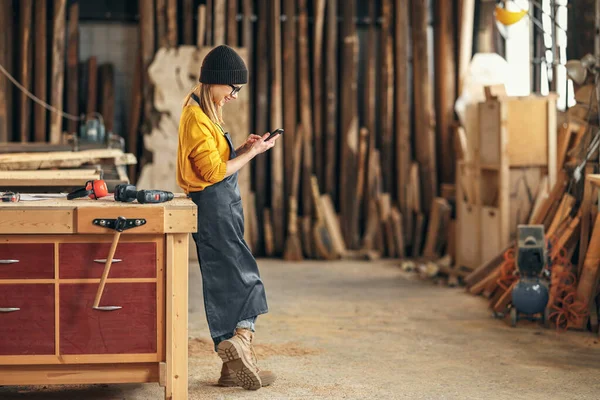 站在精品车间工作台附近的年轻女伐木工人在手机围裙信息传递方面的全景照片 — 图库照片