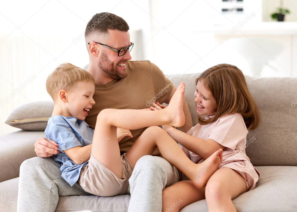 Spending weekend with dad. Young loving father playing having fun with two little kids, son and daughter while relaxing together on sofa at home. Positive happy family enjoying time together