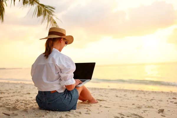 Working remotely on seashore. Young successful woman female freelancer in straw hat working on laptop while sitting on tropical beach at sunset, full length. Distance work concept