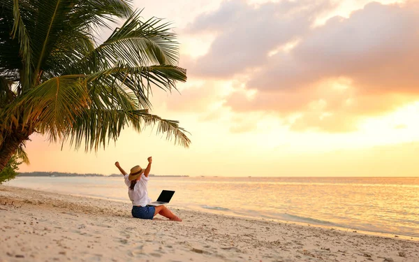 Work from anywhere. Rear view of young woman, female freelancer in straw hat working on laptop, keeping arms raised and cellebrating success while sitting on the tropical sandy beach at sunset