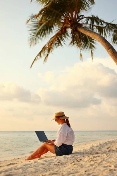 Deniz kıyısında uzaktan çalışıyor. Günbatımında tropikal plajda tam uzunlukta otururken hasır şapkalı başarılı kadın serbest yazar. Mesafe çalışma kavramı