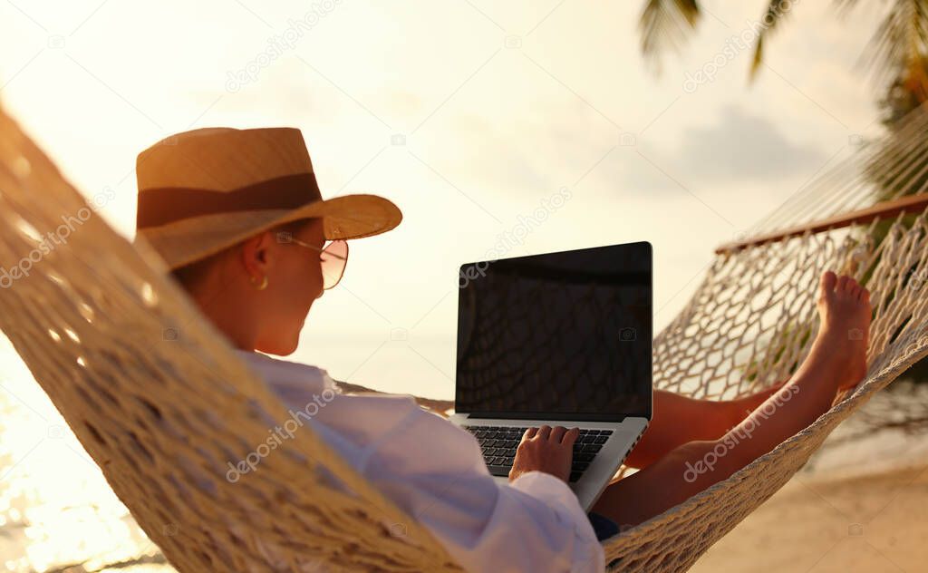 Back view of young woman, successful female freelancer using laptop while lying in hammock on the tropical beach at sunset, working remotely during summer vacation. Distance work concept