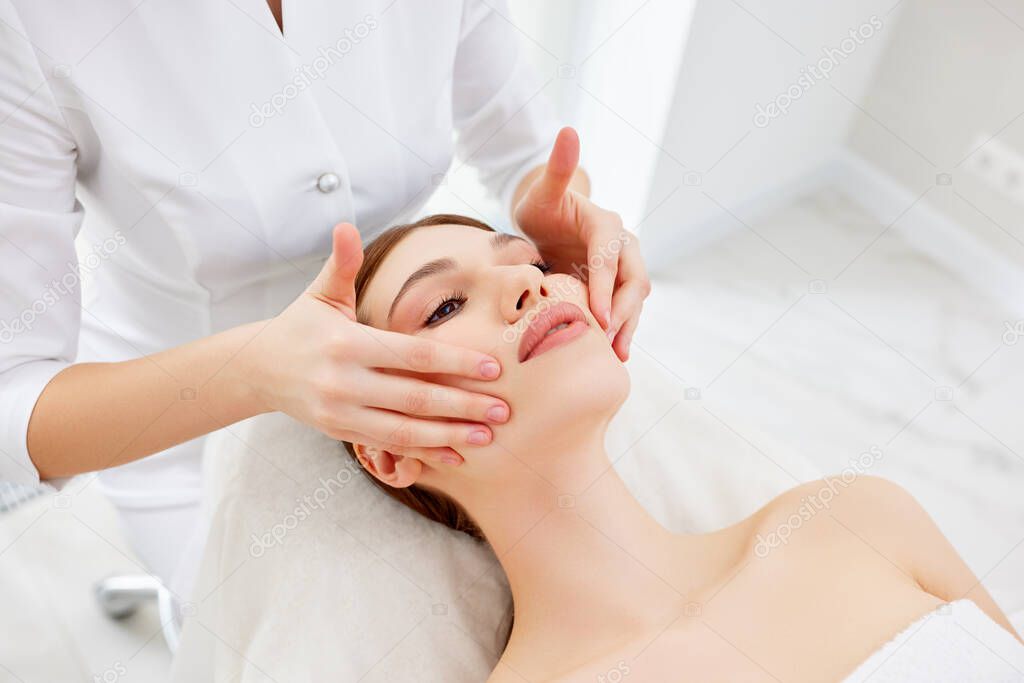 Close up of beautician or masseur doing myofascial or buccal head and face massage for young woman. Female patient lying at spa center, receiving anti aging beauty treatment. Skin rejuvenation concept