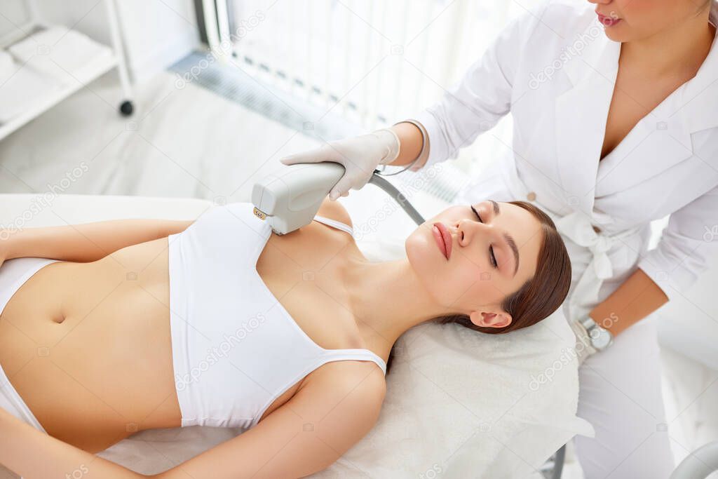 From above of young female client receiving laser procedure ultrasound skin lifting  during rejuvenation procedure in beauty clinic on neck, decollete area and chest in modern salon