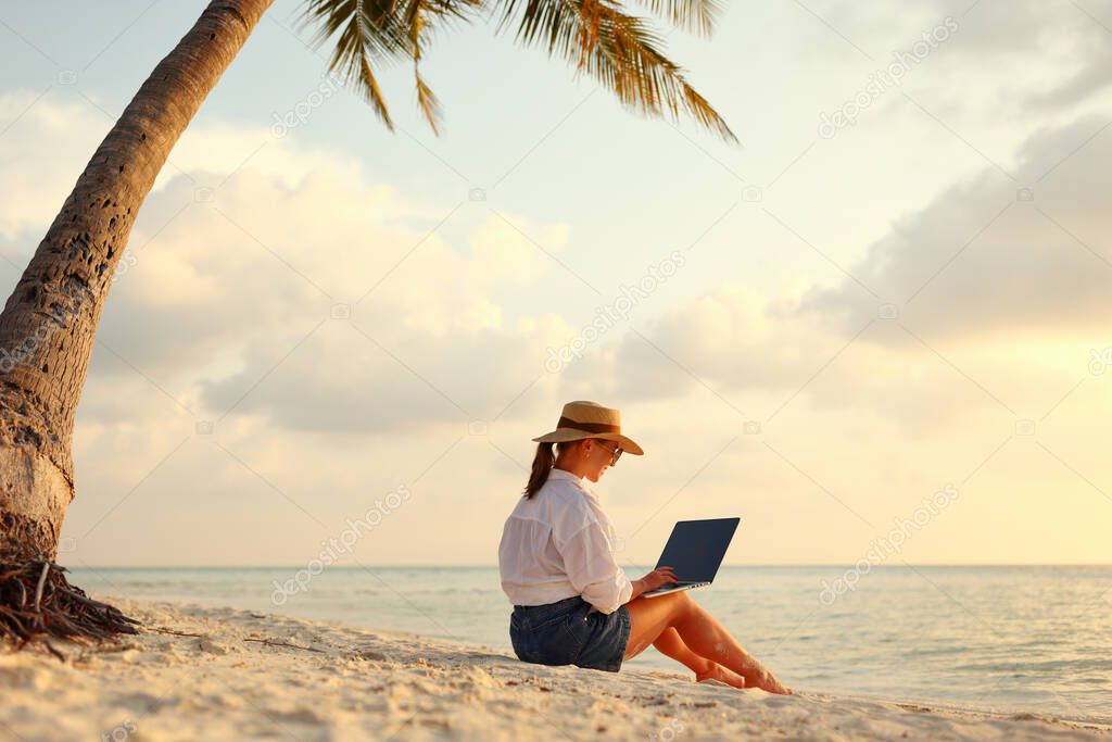 Working remotely on seashore. Young successful woman female freelancer in straw hat working on laptop while sitting on tropical beach at sunset, full length. Distance work concept