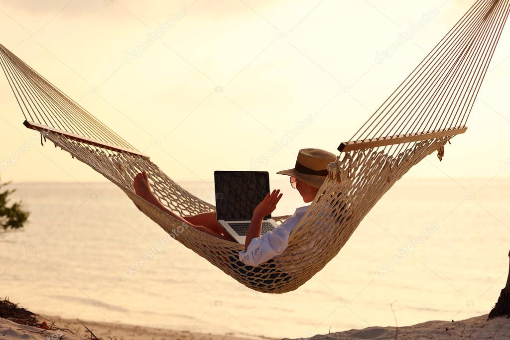 Having online conversation. Young happy successful woman wearing straw hat waving and smiling while having video call on digital laptop, relaxing in the hammock on tropical sandy beach on sunny day