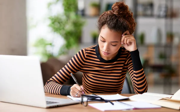 若いアフリカ系アメリカ人女性に焦点を当て 職場に座ってリモートで作業しながらノートパソコンでメモを取ります 家庭でオンライン学習の集中混合レースの女性学生 — ストック写真