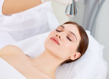Modern güzellik kliniğindeki kadın müşterinin yüzüne lazer teçhizatı kullanan profesyonel bir kozmetikçi.