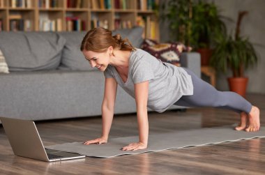 Spor kıyafetleri giyen, spor giyinen, Asya 'da oturma odasında durup dizüstü bilgisayar ekranına bakıp gülümseyen genç ve mutlu bir kadın. Covid19 salgını sırasında spor