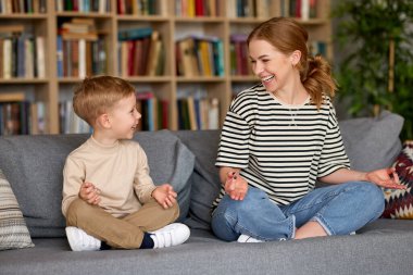Çocuklu yoga. Genç, pozitif bir anne, oğlana nilüfer çiçeğinin üzerinde birlikte otururken nasıl meditasyon yapılacağını öğretirken, evdeki oturma odasında, aktif anne ve çocuk, aile aktivitelerinin keyfini çıkarıyorlar.