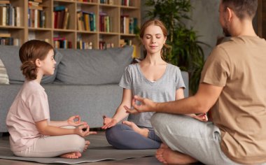 Genç ebeveynler evde çocukla meditasyon yapıyor. Güzel bir aile babası, annesi ve çocuğu. Gözleri kapalı. Lotus poz verirken yoga yapıyorlar.