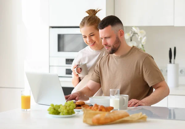 朝一緒に朝食を楽しみながらノートパソコンの画面を見ている若い美しい幸せな家族のカップルは 白いモダンなキッチンに立っています 妻と夫は食事をしながらオンラインでニュースを読む — ストック写真