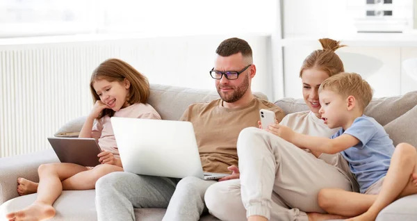 年轻快乐的家庭 有两个孩子的父母在使用现代技术的同时一起呆在家里 带着笔记本电脑 数码平板电脑和智能手机的父亲 母亲和孩子们在沙发上放松一下 — 图库照片
