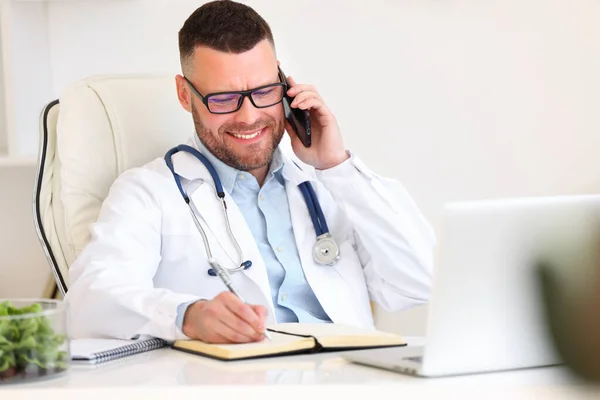スマートフォンで話してノートを作る幸せな男医師 電話で患者をコンサルティング プロの男性医師療法士ホワイトコートで聴診器と肩の上に働く現代のクリニック — ストック写真