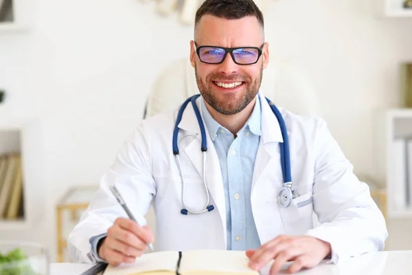 快乐友善的男医生 身穿白色外套 肩带听诊器 一边在医院工作一边微笑着看着相机 一边坐在工作场所等待病人 — 图库照片