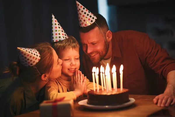 家で小さな息子の誕生日を祝うパーティー帽子をかぶっている若い幸せな家族 かわいいです男の子で目を閉じて願いを作り 暗い部屋でチョコレートケーキの上にろうそくを吹きます — ストック写真