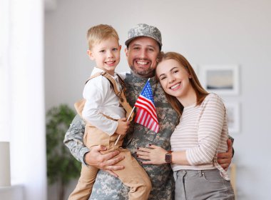 Happy young man father in military uniform reunited with his beautiful american family, hugging with smiling wife and cute kid son with flag of United States in hand, meeting daddy from US army at home