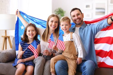 Genç mutlu Amerikalı aile babaları ve iki küçük çocuk evde kanepede oturmuş Amerika Birleşik Devletleri bayraklarıyla kameralara gülümsüyor ve Bağımsızlık Günü 'nü kutluyorlar. Vatansever Amerikan bayramı konsepti