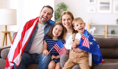Genç mutlu Amerikalı aile babaları ve iki küçük çocuk evde kanepede oturmuş Amerika Birleşik Devletleri bayraklarıyla kameralara gülümsüyor ve Bağımsızlık Günü 'nü kutluyorlar. Vatansever Amerikan bayramı konsepti