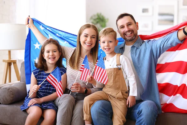 若い幸せなアメリカの家族の両親と2人の小さな子供たちは 独立記念日を祝いながら 米国の国旗とカメラで笑顔で自宅でソファに座っています 愛国的なアメリカの休日の概念 — ストック写真