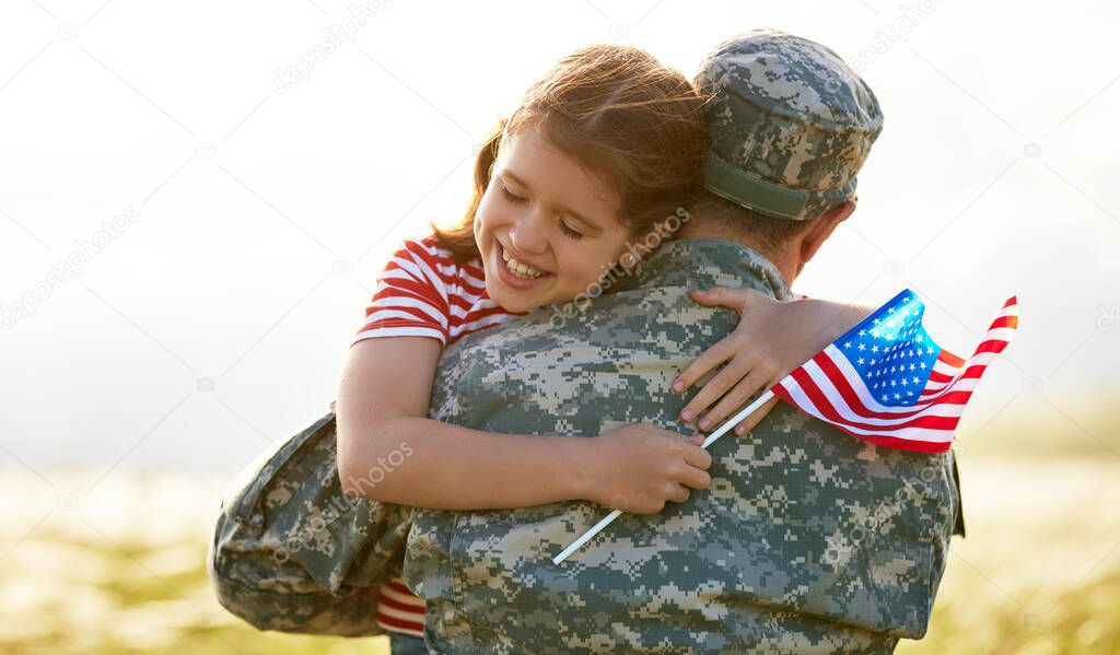 Happy little girl daughter with American flag hugging father in military uniform came back from US army, rear view of male soldier reunited with family while standing in green meadow on summer day