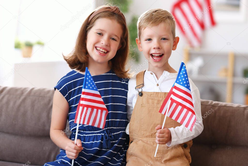 Two cute little children brother and sister with USA flags smiling at camera while celebrating Independence day of United States on July 4 with family at home, sitting on sofa in modern apartment